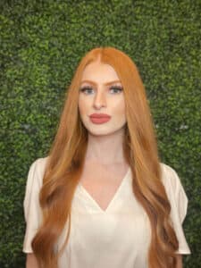 A woman with long red hair, blue eyes, rose colored lipstick and a cream colored shirt in front a a green hedge.