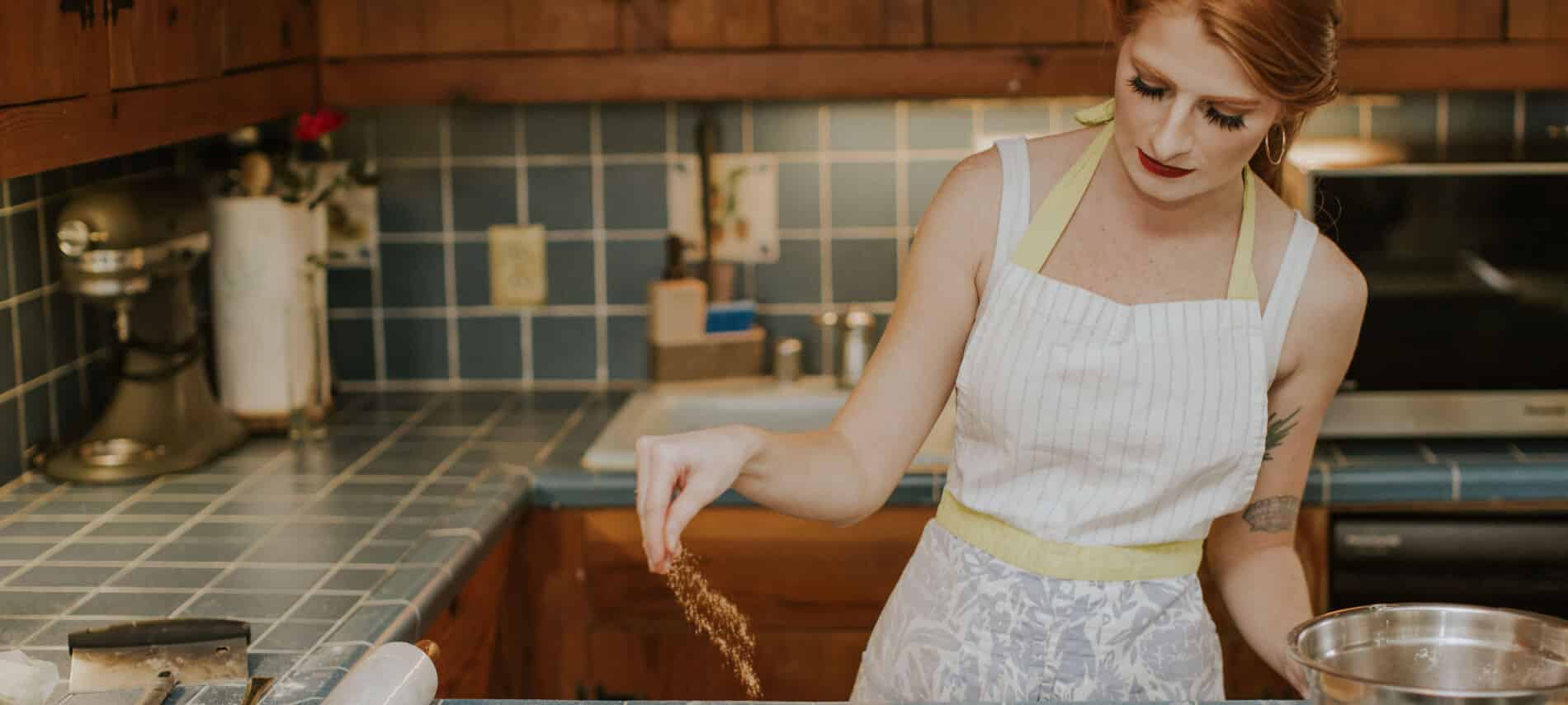 A woman with red hair wearing an apron in a kitchen sprinkling cinnamon.