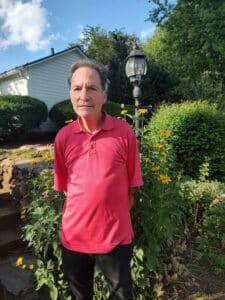 A man with a red shirt and black pants, his hands behind his back standing in front some bushes with yellow flowers, and a white house in the background.