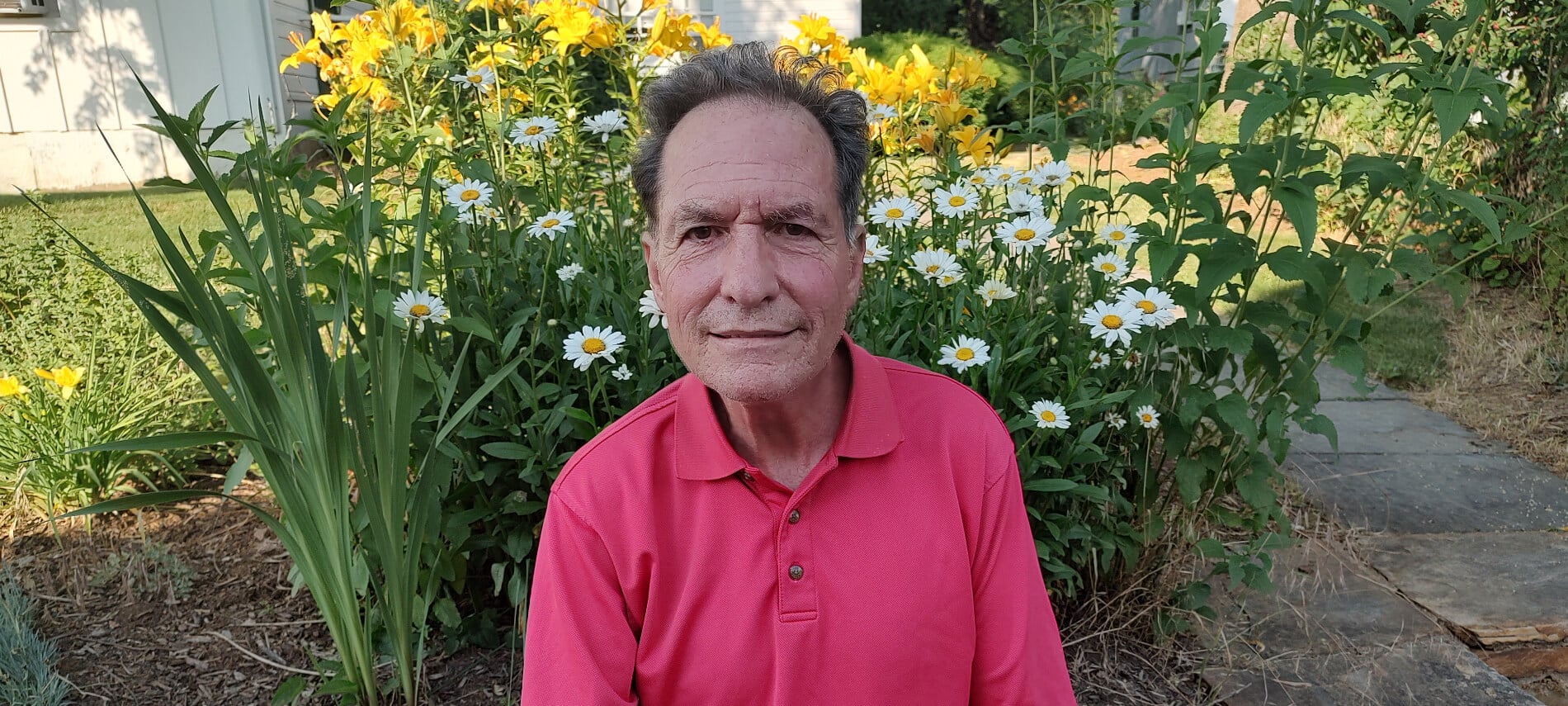 A man in a red shirt with a slight smile outside in front of some yellow and white flowers.