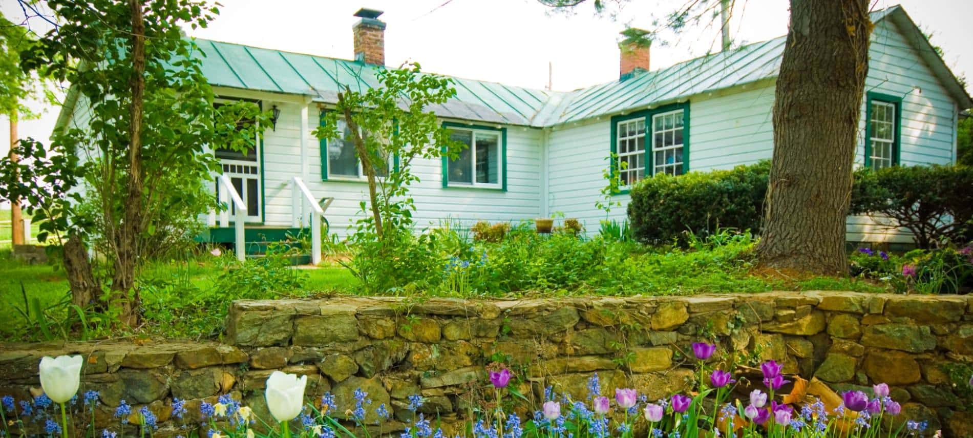 Exterior view of cottage painted white with green trim surrounded by green grass and shrubs, a stone retaining wall, and white, purple, and blue flowers