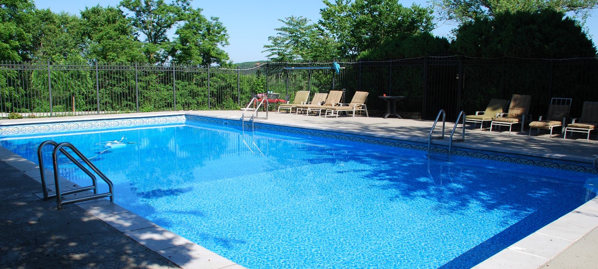 Large pool with blue and white tile surrounded by rod iron fence and large trees with green leaves