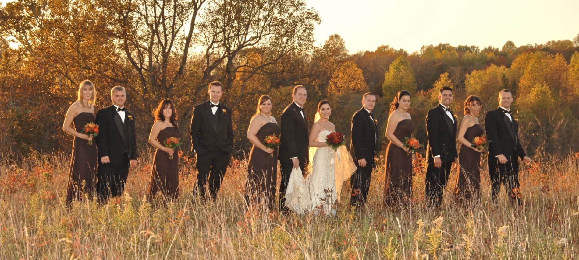 Large wedding party with bride in cream, bridesmaids in dark brown, and groom and groomsmen in black suits standing on hill in front of trees with fall color