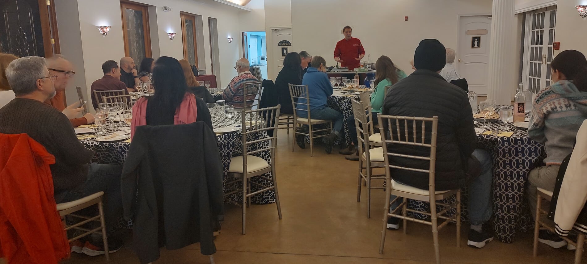 A man in a red shirt at the front of a dining room teaching a cooking lesson to a group of people seated at tables.